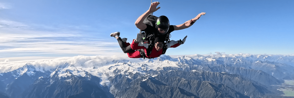 Two people tandem skydiving over snow-capped mountains.
