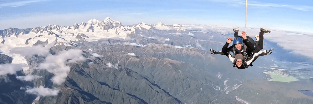 Tandem skydivers above mountains and clouds.