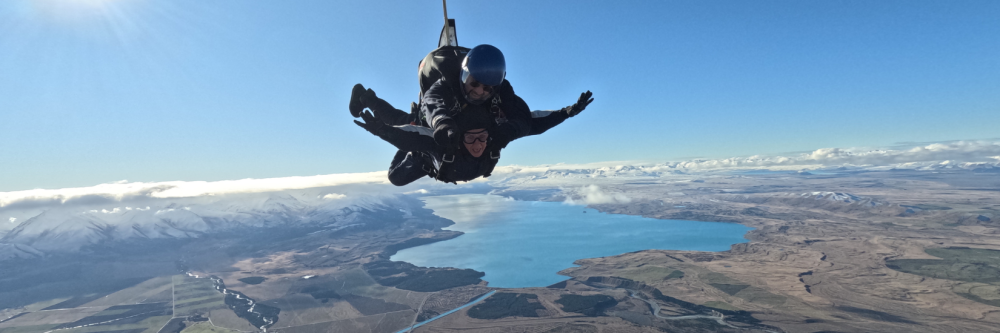 Two skydivers in freefall above a large lake and mountainous landscape.