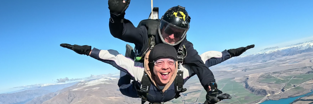 Two people tandem skydiving with mountains and a lake below.