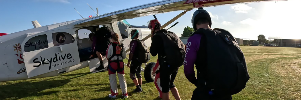 Skydivers board a plane on grassy field with clear sky.