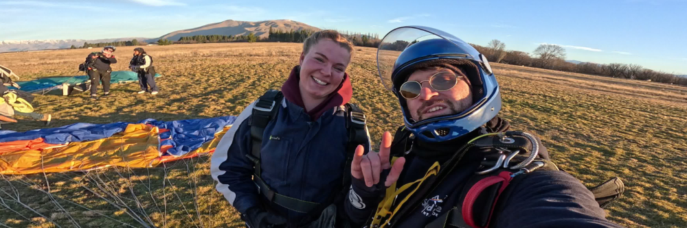 Two people in skydiving gear smiling on a grassy field with parachutes around.