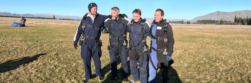 Four people in jumpsuits smiling on a grassy field after skydiving.