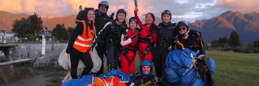 Group of skydivers in gear posing happily on grass with mountain backdrop.