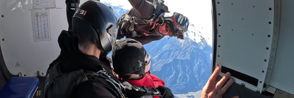 Skydivers exiting a plane with mountain view in the background.