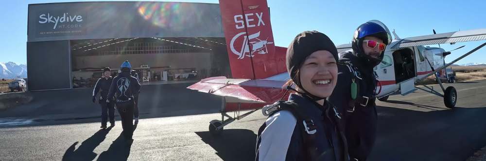 Smiling people in skydiving gear near a small plane and hangar.