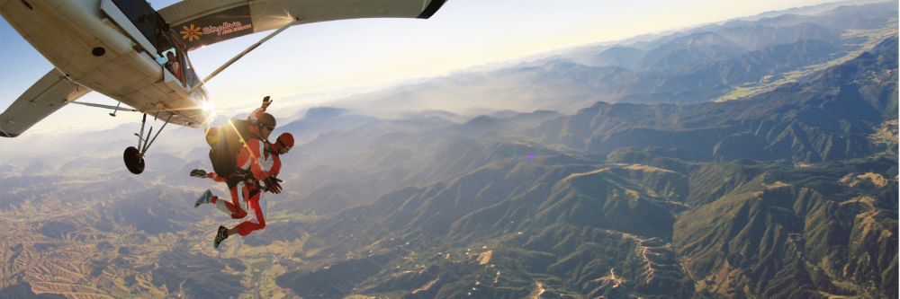 Tandem skydivers jump from a plane over a vast mountainous landscape.