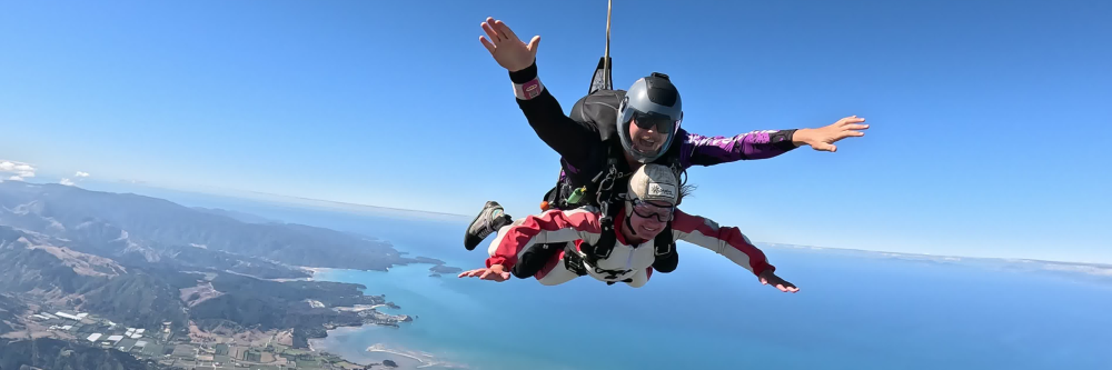 Two people tandem skydiving above coastal landscape.