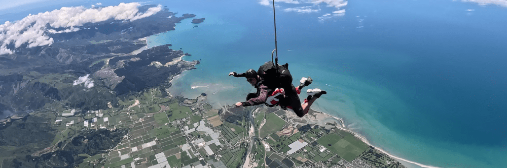 Two people tandem skydiving over a coastal landscape with fields and ocean views.