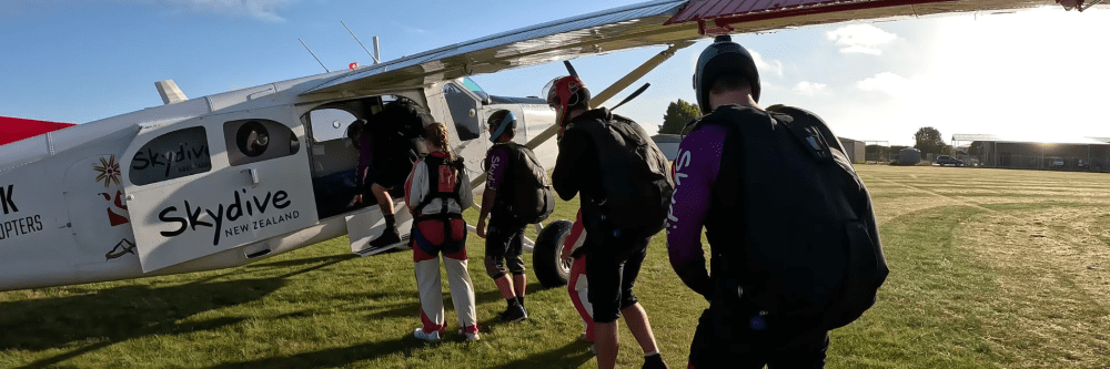 Four skydivers board a small plane with 'Skydive' logo on a sunny day.