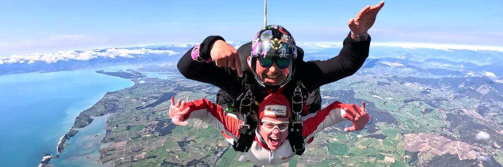 Two people tandem skydiving over a scenic landscape with a large body of water.
