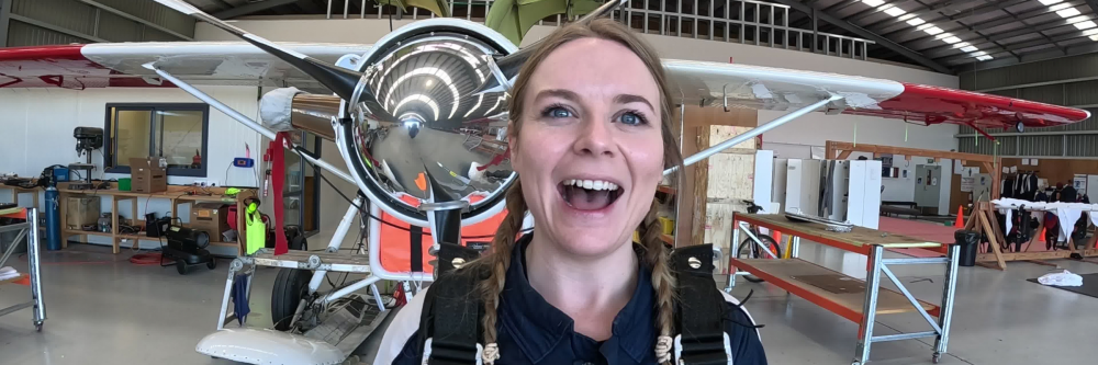 Person in parachute gear smiling in front of a small aircraft in a hangar.