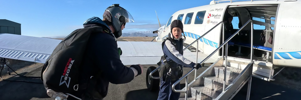 Two skydivers board a small airplane, one on stairs, one on the ground, with clear skies in the background.