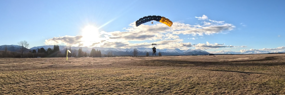 Parachutist landing in a field with mountains and sun in the background.