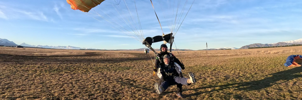 Two people tandem paragliding, landing on a grassy field.
