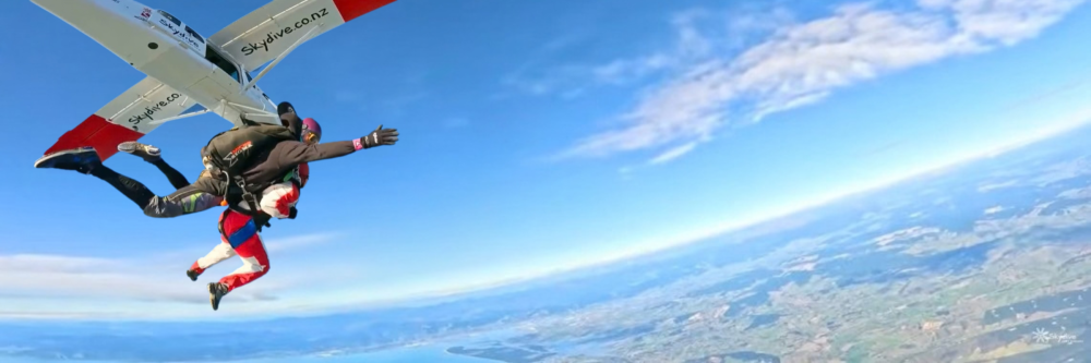 Skydivers jumping from a plane above scenic landscape, with lake and hills.