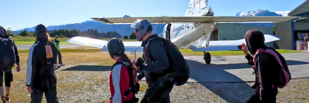 People in skydiving gear walk towards a small airplane on a clear day.