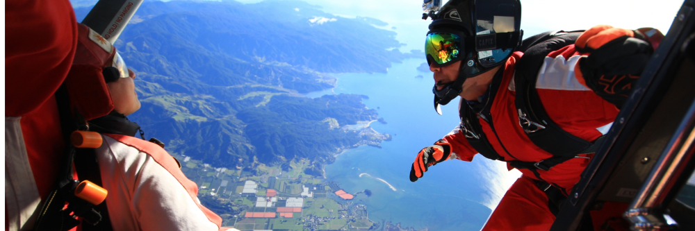 Two skydivers in orange suits prepare to jump from a plane over a scenic landscape.