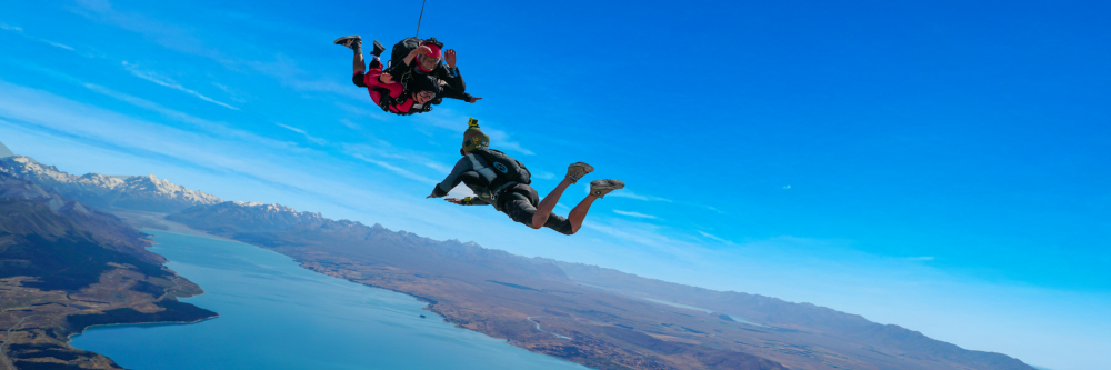 Two people skydiving over a vast landscape with a lake and mountains.