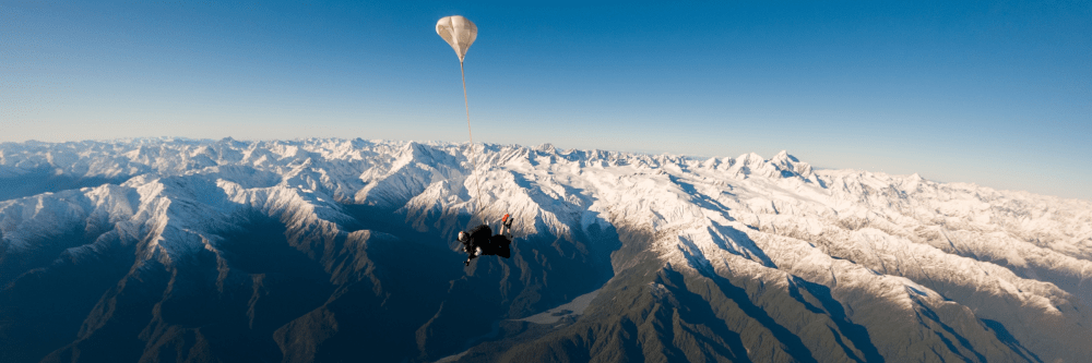Parachuter above snow-covered mountains under a clear blue sky.