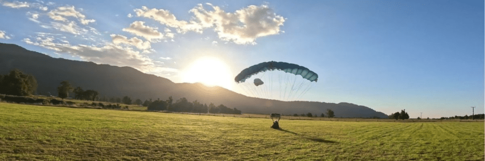 Paraglider landing on grassy field at sunset, mountains in background.