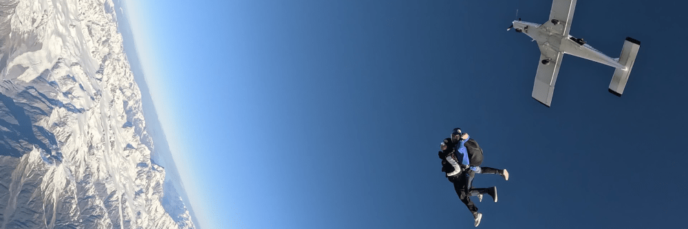 Two skydivers jump from a plane, snow-capped mountains below.