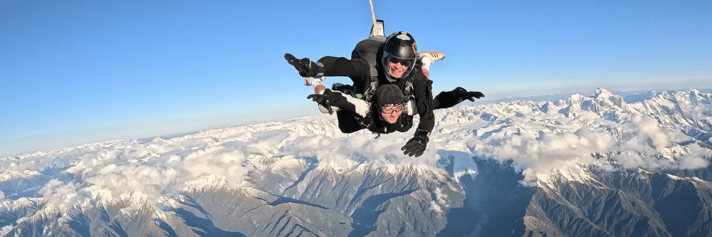 Two skydivers in freefall above snow-covered mountains and clouds.