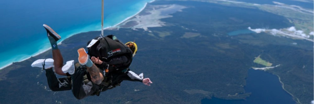Two people tandem skydiving over coastal landscape with ocean view.